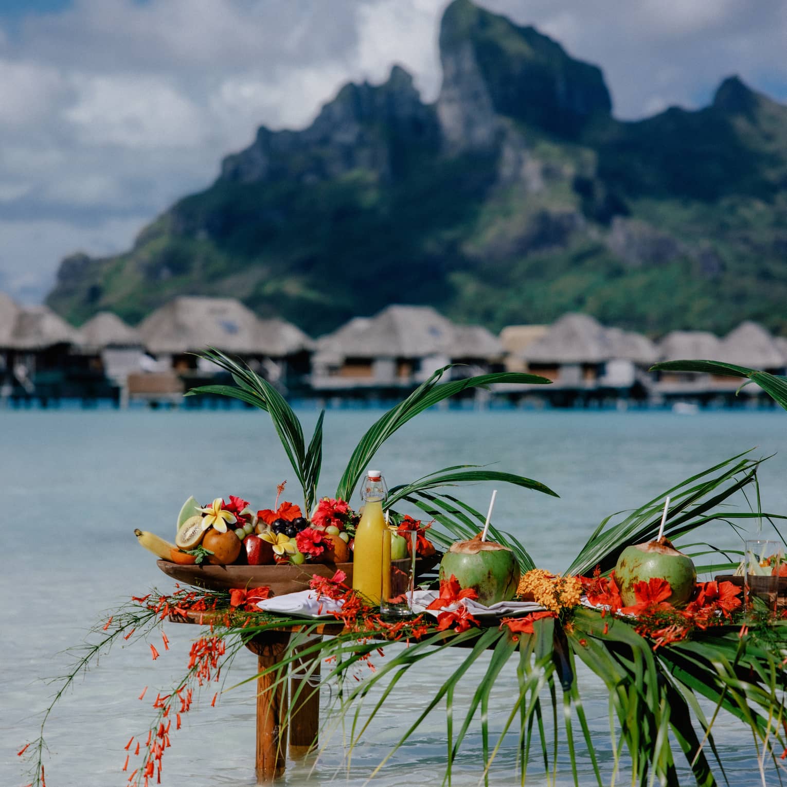 Small wooden table with legs submerged in clear water, topped with assorted fruit and coconuts with straws, mountain and overwater pavilions in background