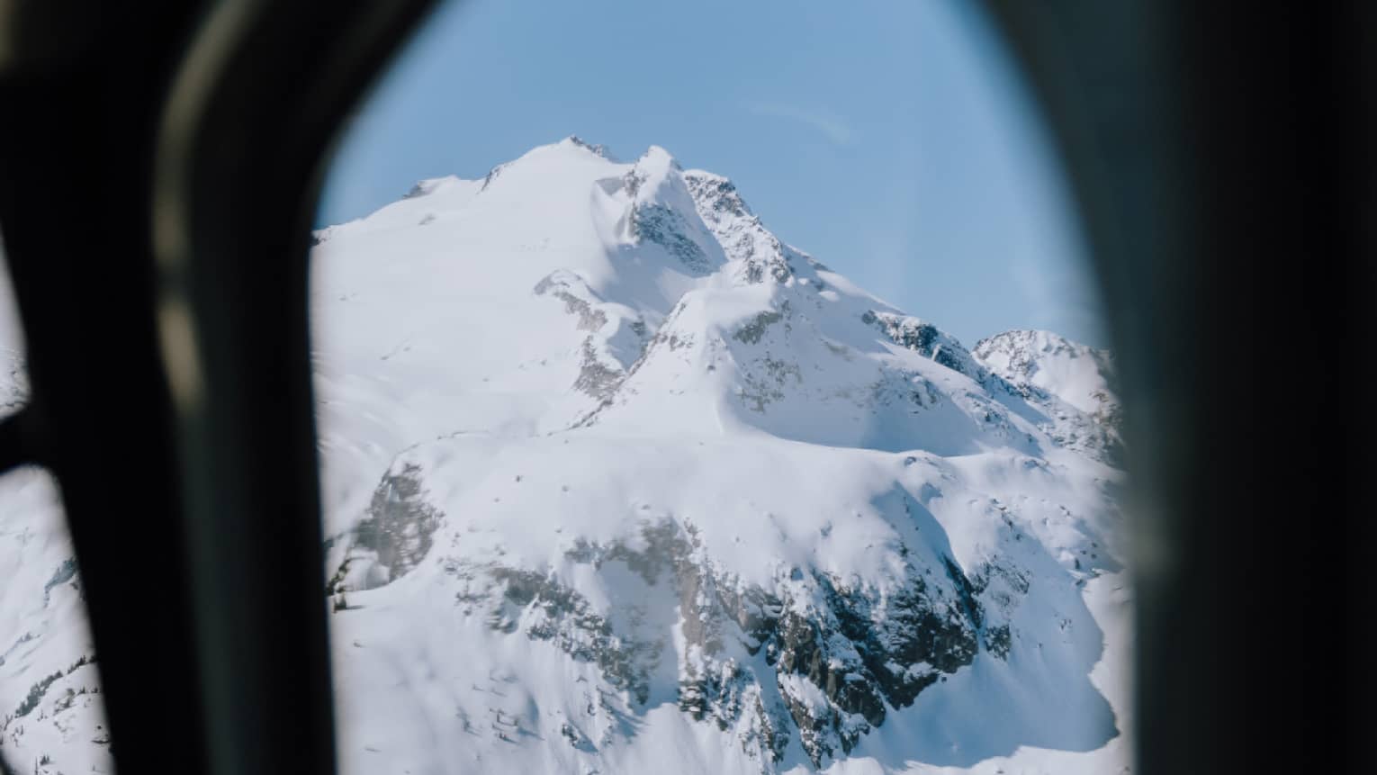 An aerial view through an airplane window of a mountain covered in snow with sporadic clumps of trees around the base.