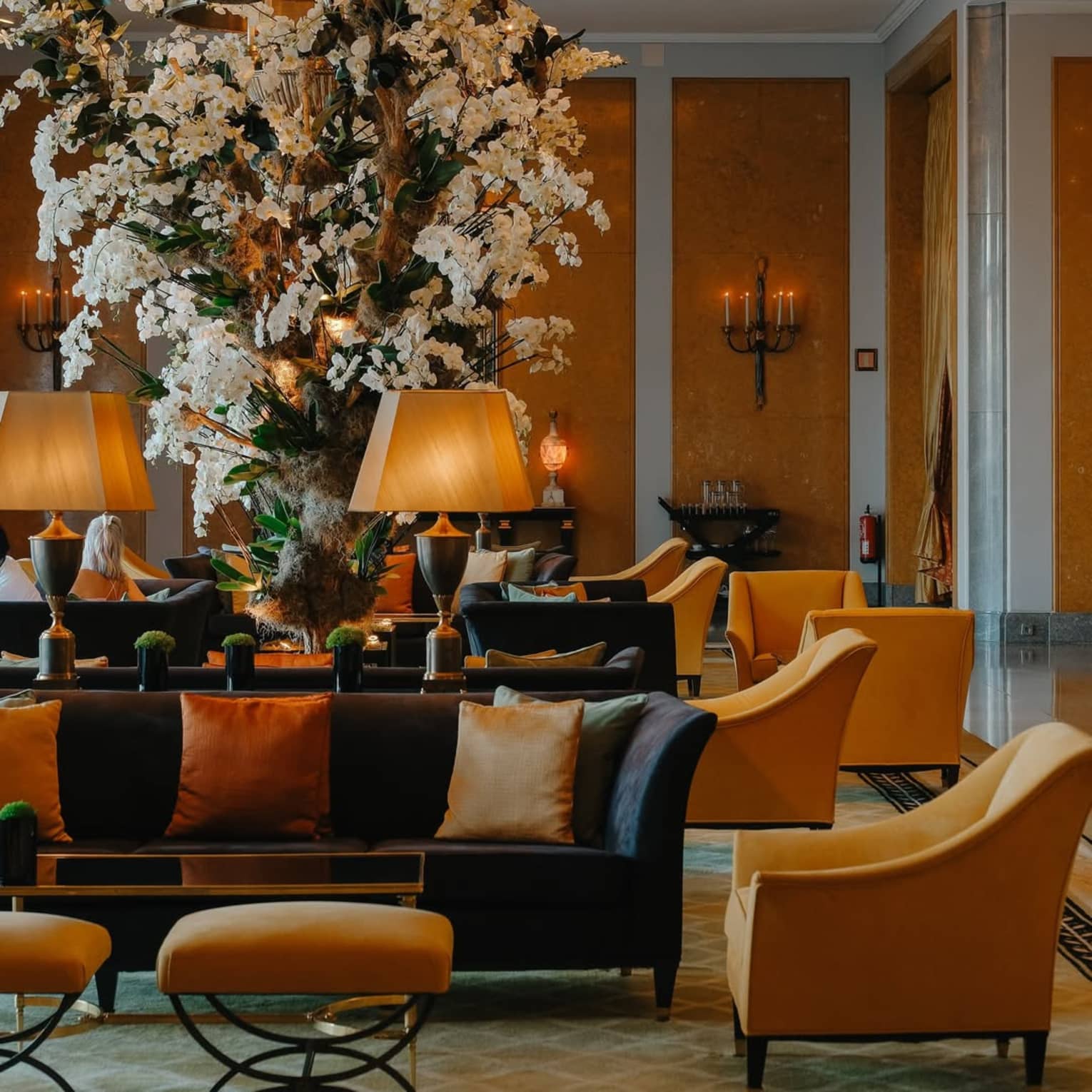 A spacious hotel lobby with dark grey couches, yellow upholstered chairs, a chandelier and a huge white floral arrangement that touches the ceiling