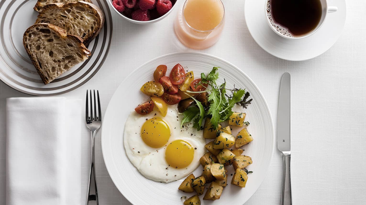 Breakfast plate with sunny-side-up eggs, cherry tomatoes, greens, and roasted potatoes, alongside toast, raspberries, grapefruit juice and coffee.