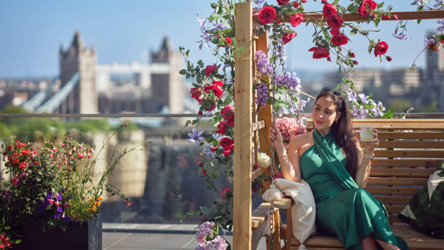 Woman in green dress sitting on terrace bench under arbor of red roses with Tower Bridge in backdrop
