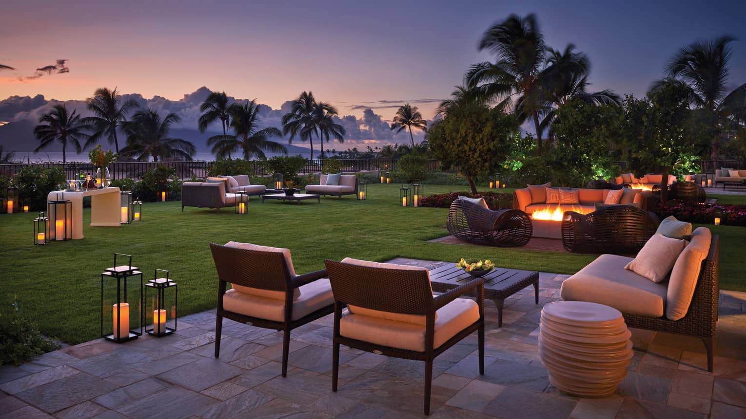 Outdoor lounge area at sunset with wicker seating, fire pits, palm trees and lantern lighting