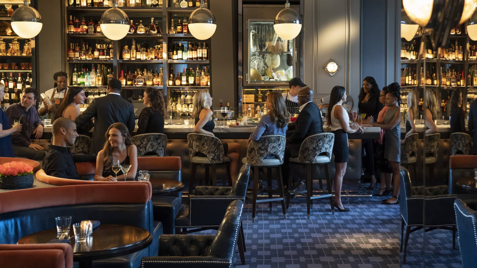 Guests and staff in a spacious, softly lit lounge space with both banquette and bar seating, hanging globe lights, and walls of shelves with bottles.