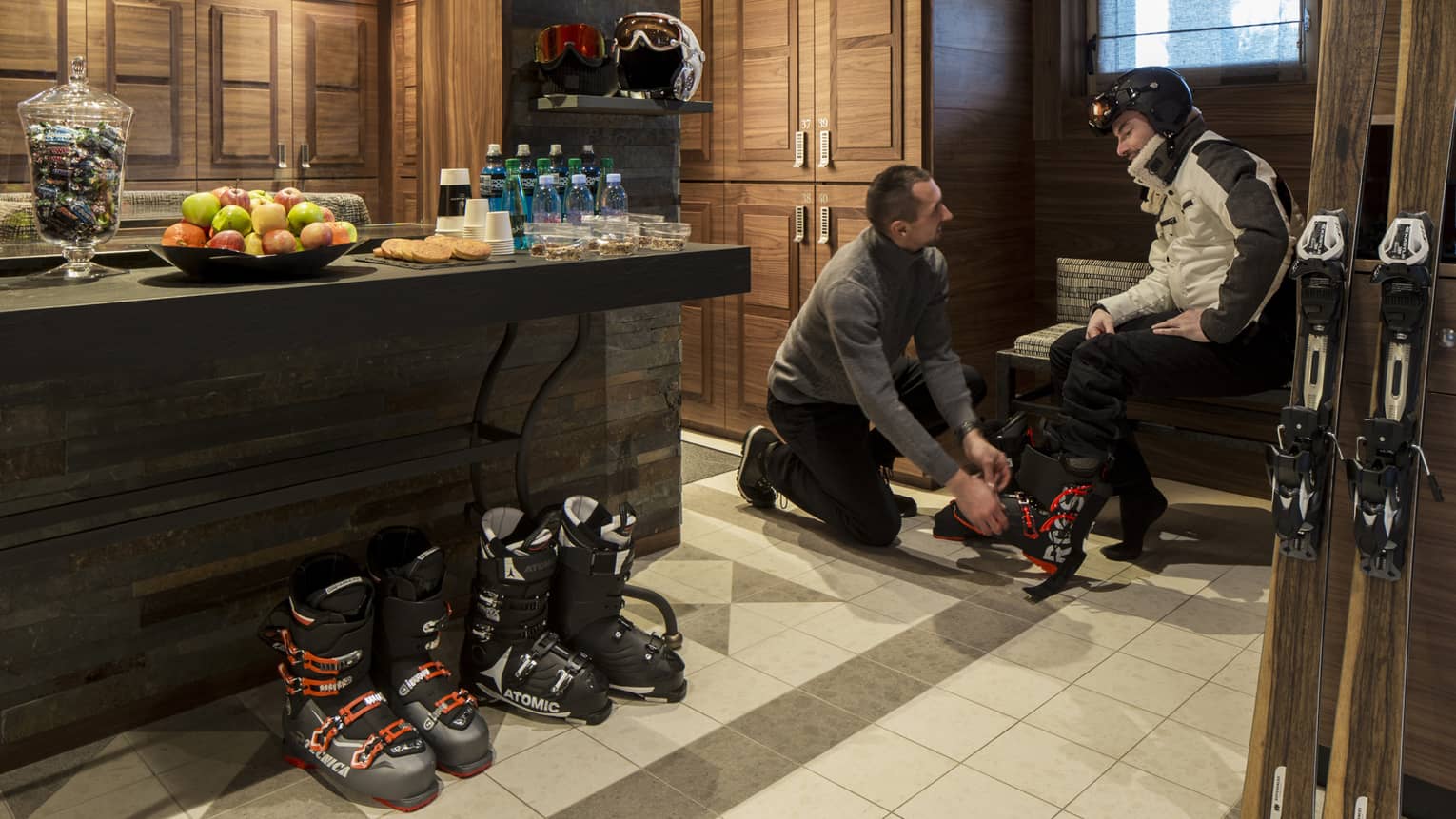 Ski concierge fits boots on man sitting on bench by skis, table with fresh fruit water