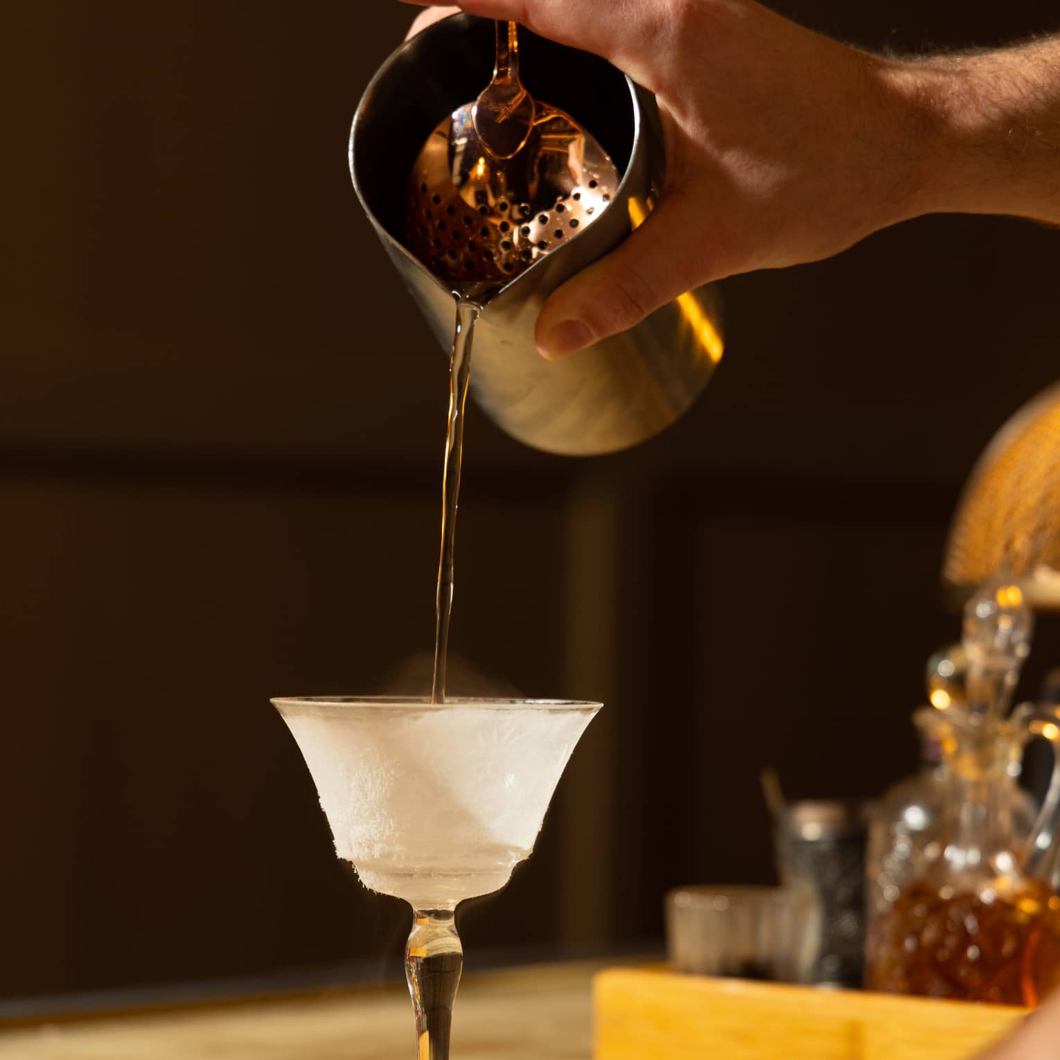 A cocktail being poured into a glass in a dimly lit bar area.