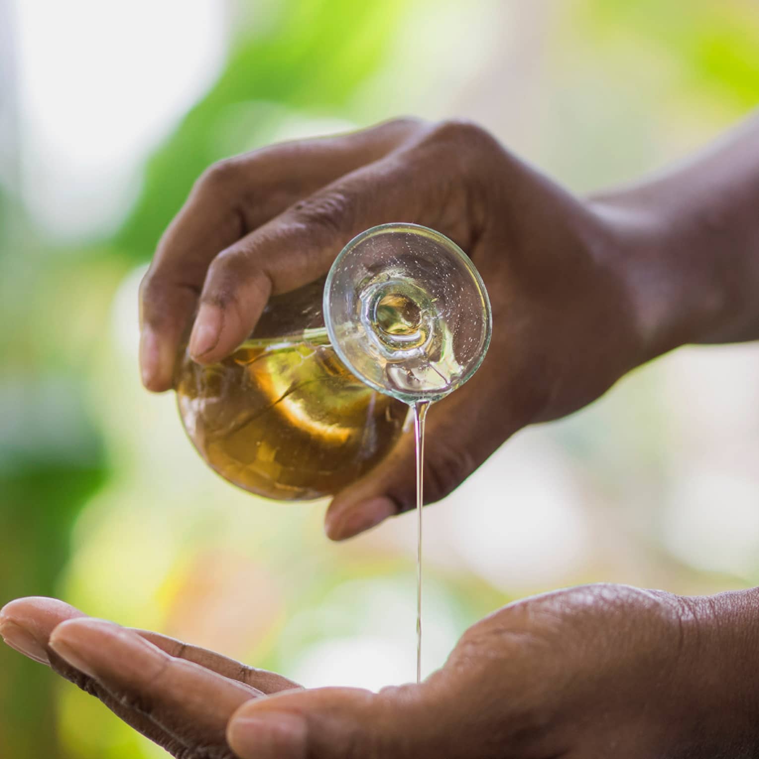 Golden oil flows from a small carafe held in one hand onto the other, a small wooden bowl underneath to catch the overflow.