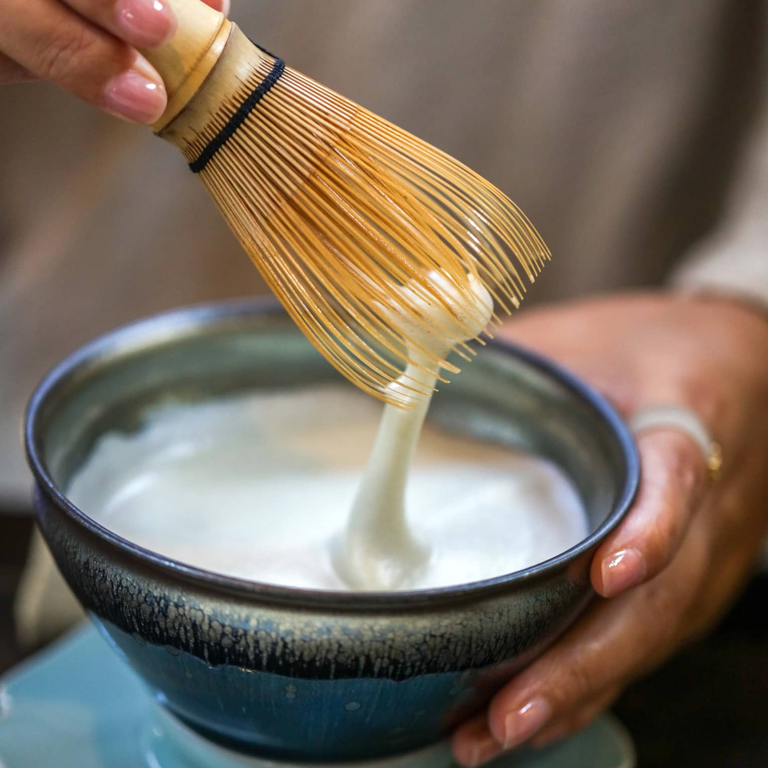 A white foamy liquid being stirred with a straw whisk in a small blue stoneware bowl on a petal-shaped base.