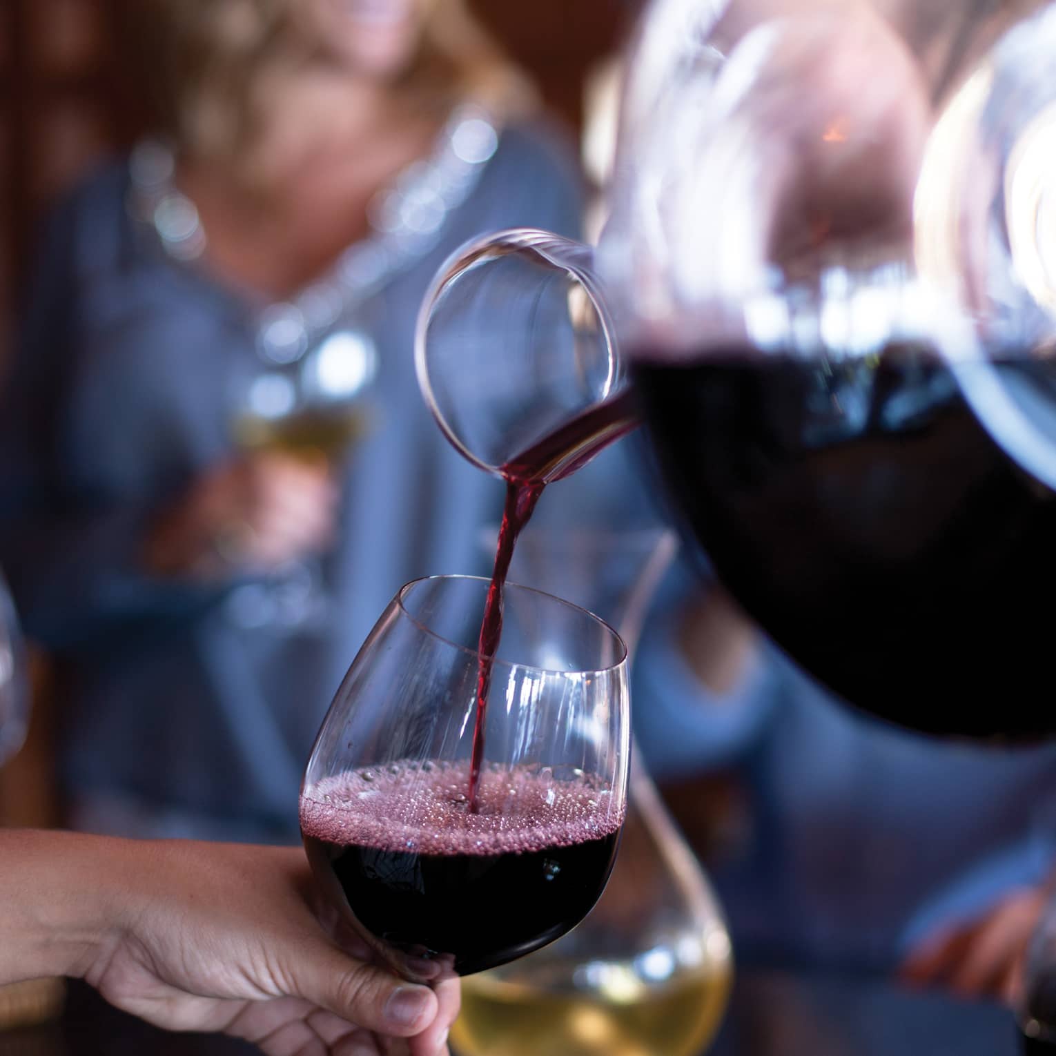 Crimson-coloured red wine is poured from a round decanter into a wine goblet, froth collecting at the surface of the glass.