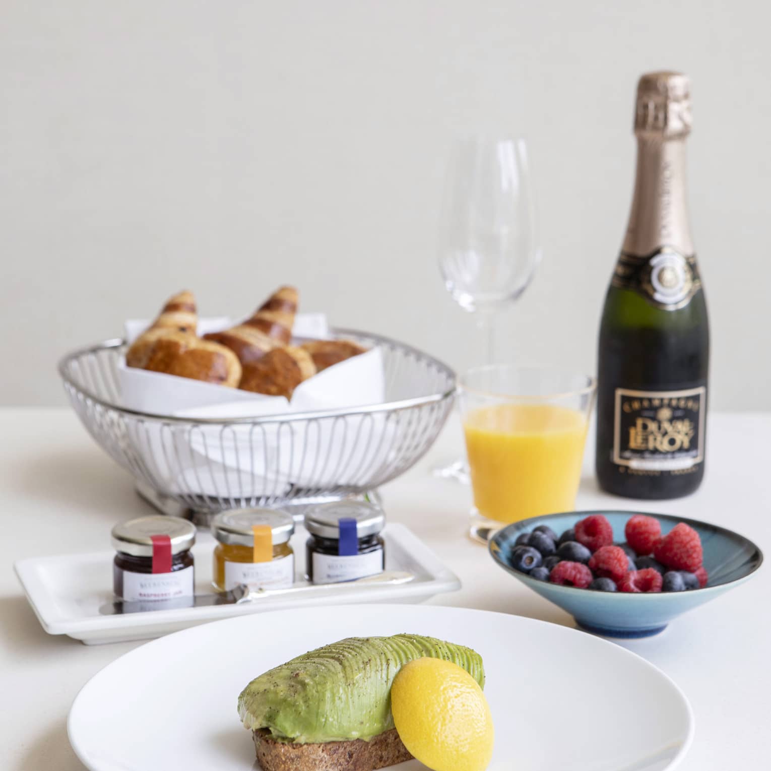 Luxurious breakfast including avocado toast, a basket of croissants, fresh fruit, and champagne and orange juice.