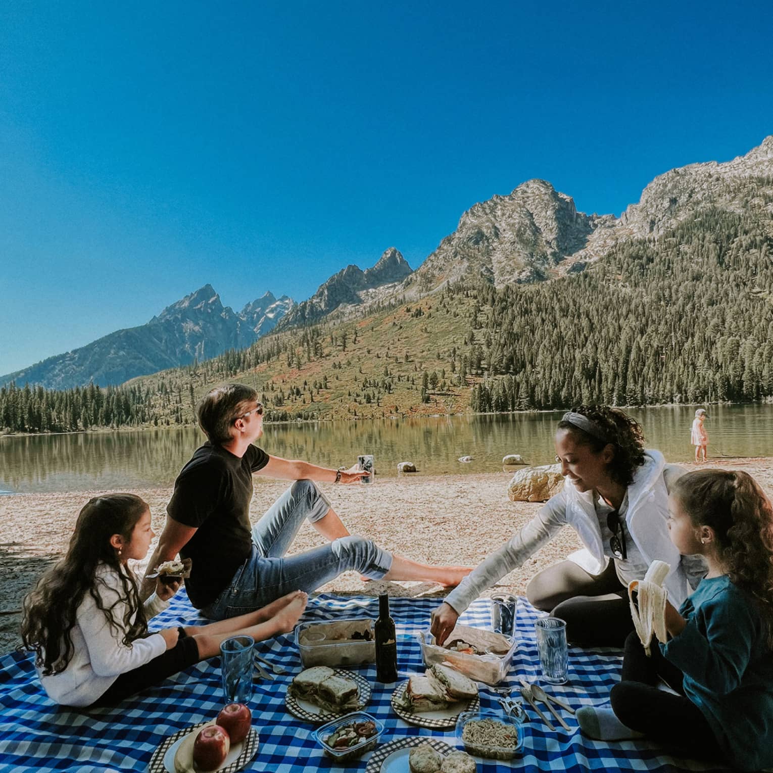 A family picnics on a checkered blanket near an alpine lake reflecting a rust and gold treed hill, distant mountains beyond,.
