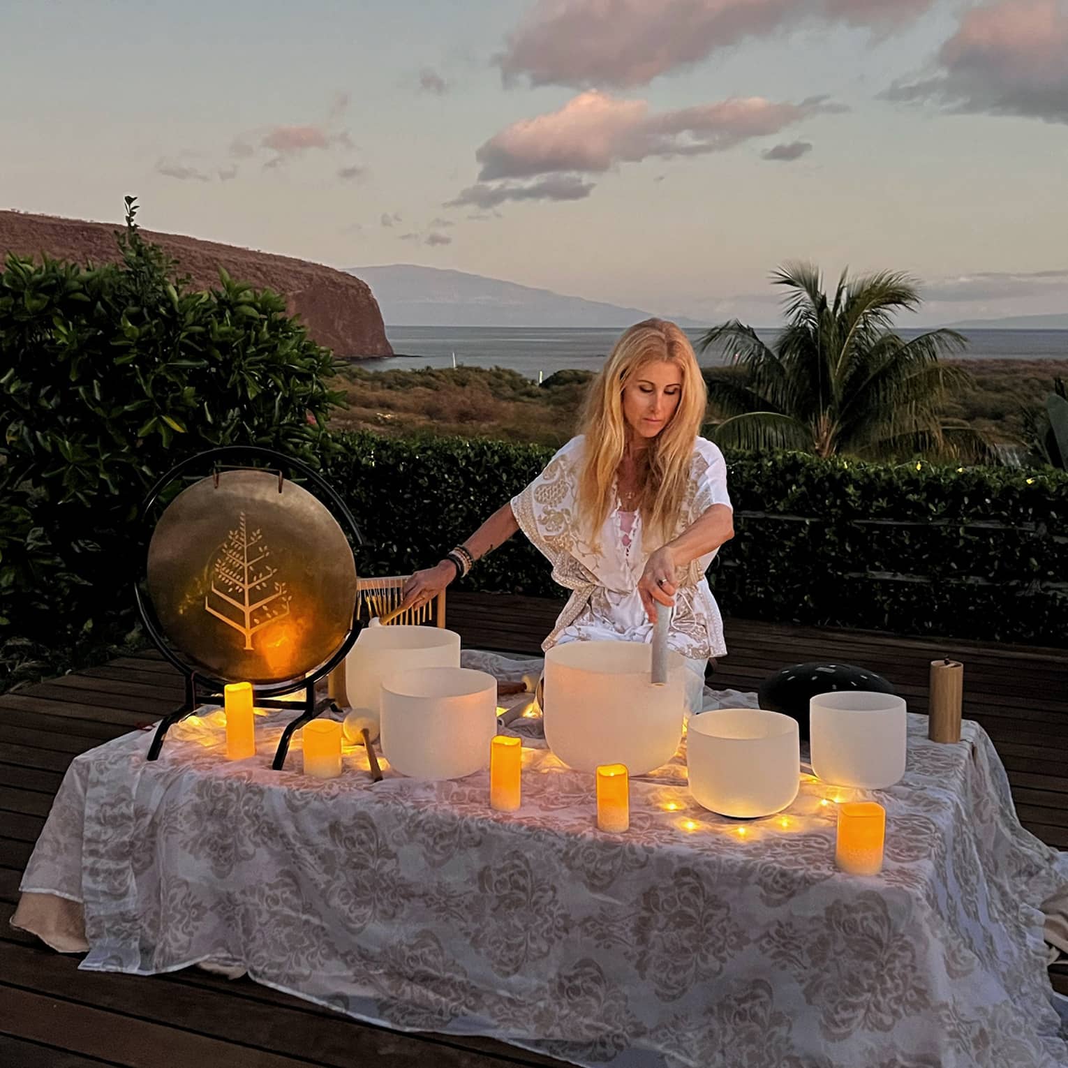 A woman performs crystal singing bowl ritual outside in beach setting at dusk