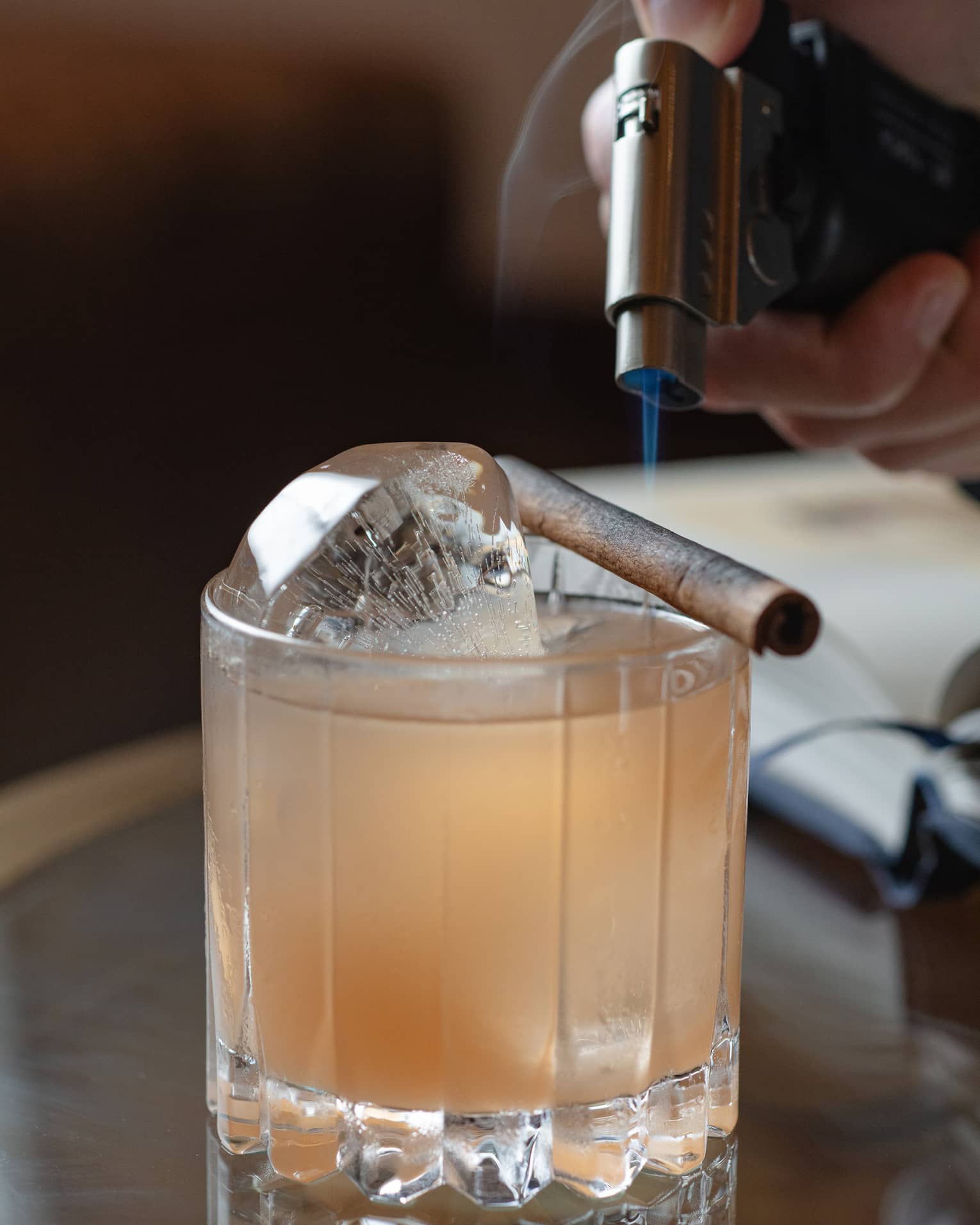 A hand holds a small blowtorch over a cinnamon stick set across the rim of a bowler glass of light amber liquid on ice.