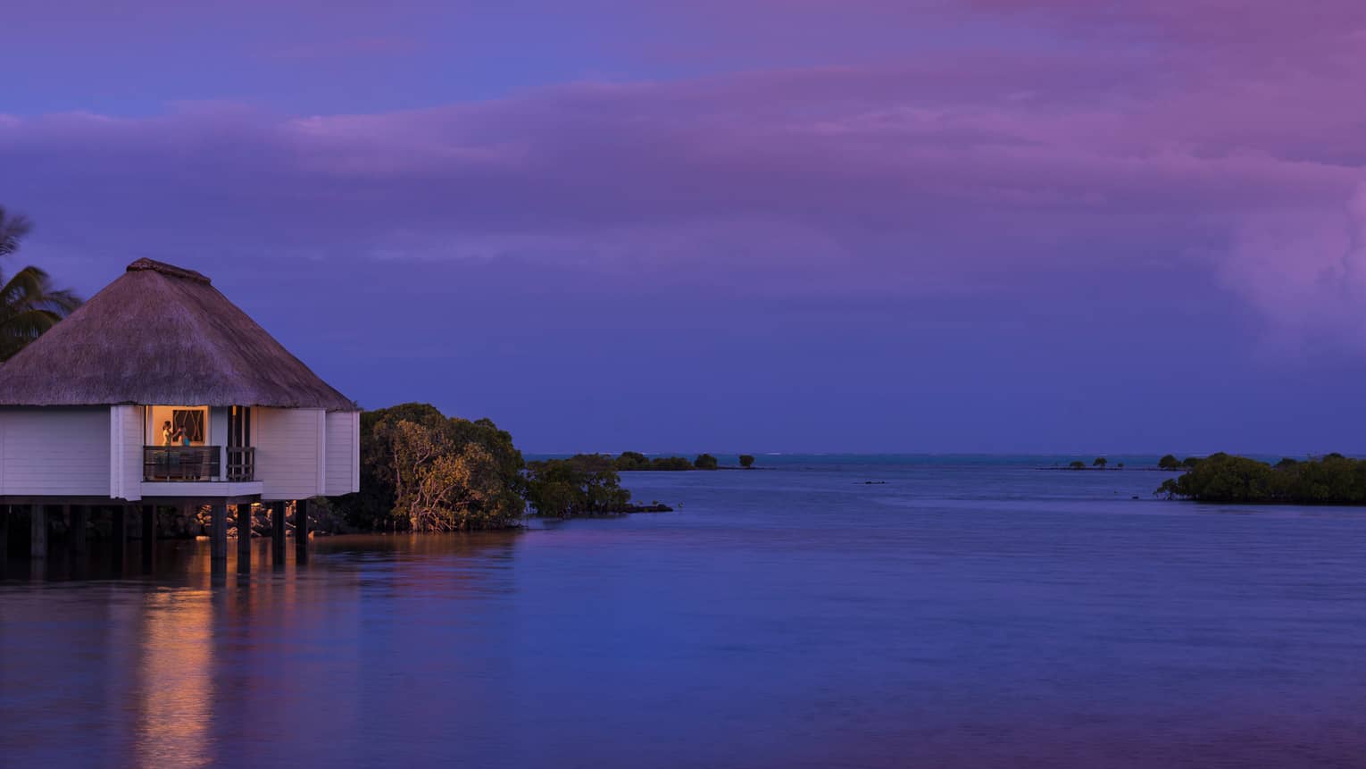 Spa staff massaging woman's head on deck of small white villa over water at night
