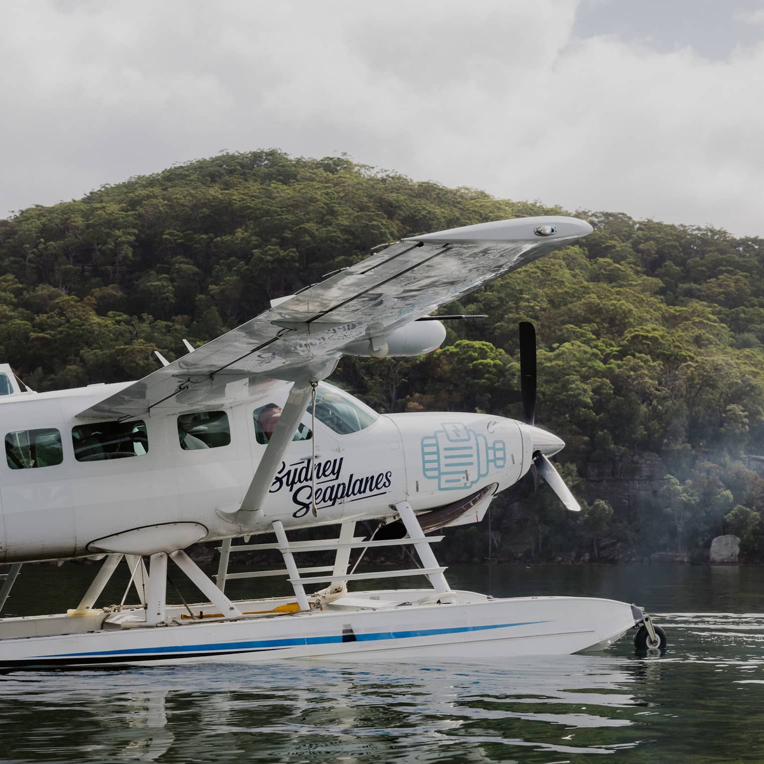 A white seaplane with the words "Sydney Seaplanes" in blue on its side, sitting on water with a lush treed hill behind it.