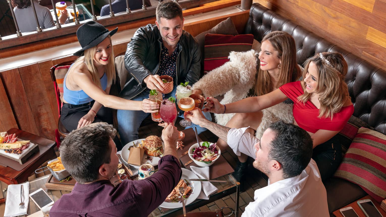 Group of friends sitting in restaurant lounge booth at table with appetizers, toast with cocktails