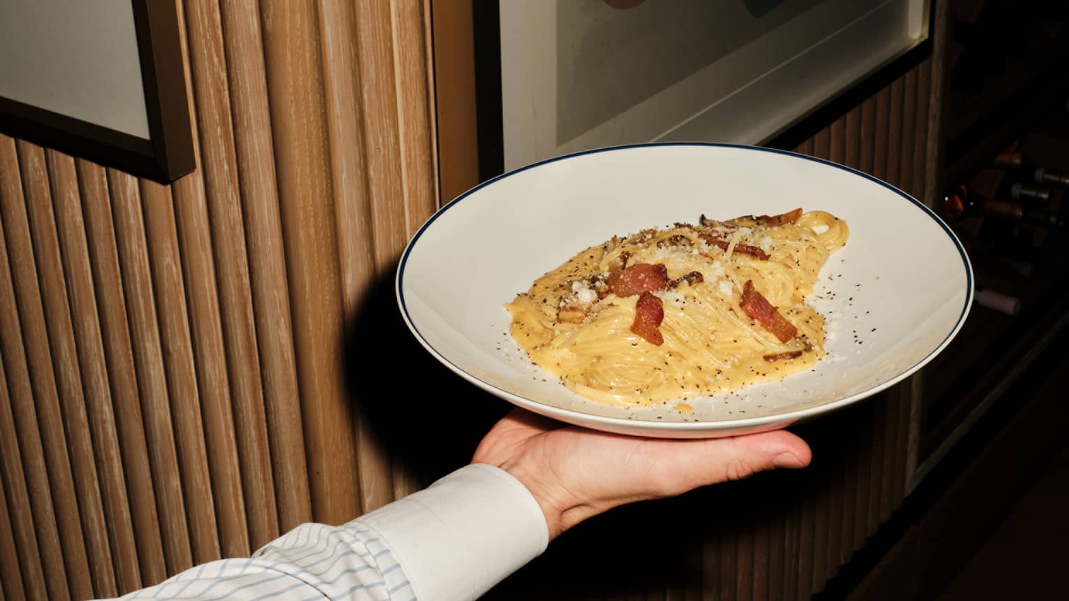 Hand holding out a plate of spaghetti carbonara topped with bacon lardons