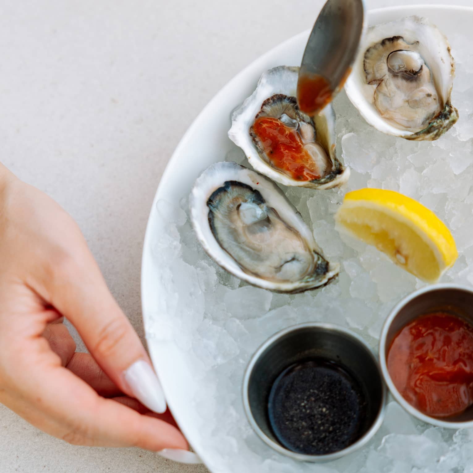 A manicured hand by a bowl of shucked oysters drizzled with red sauce, a lemon wedge and sauce cups on a bed of crushed ice.
