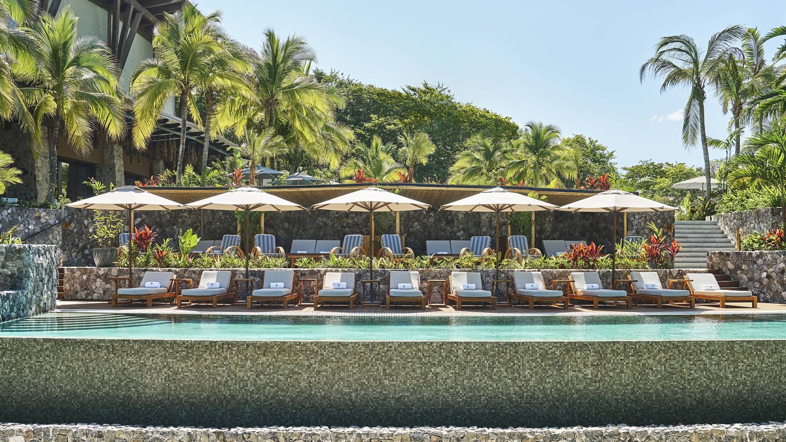 A line of lounge chairs and umbrellas at an outdoor pool surrounded by lush greenery
