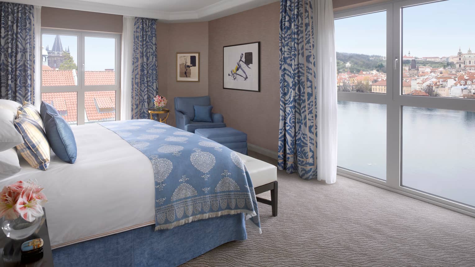 Premier Suite bed with blue blanket, pillow accents, in front of floor-to-ceiling corner windows overlooking Vltava River 
