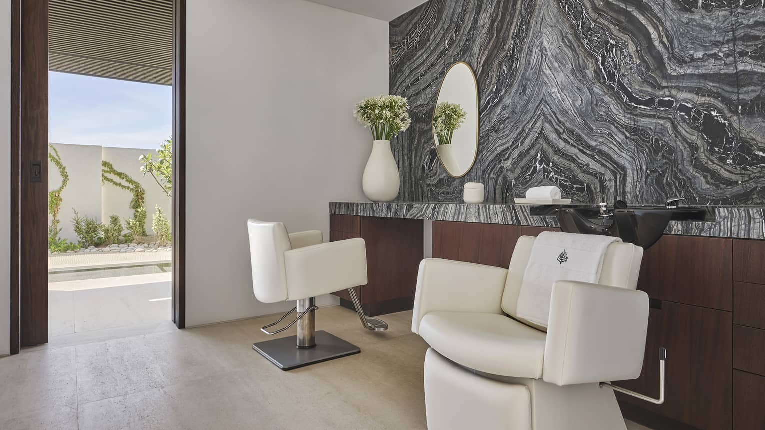 two chairs in a room with a large shelf and grey wall, a door looking at plants outside is open.