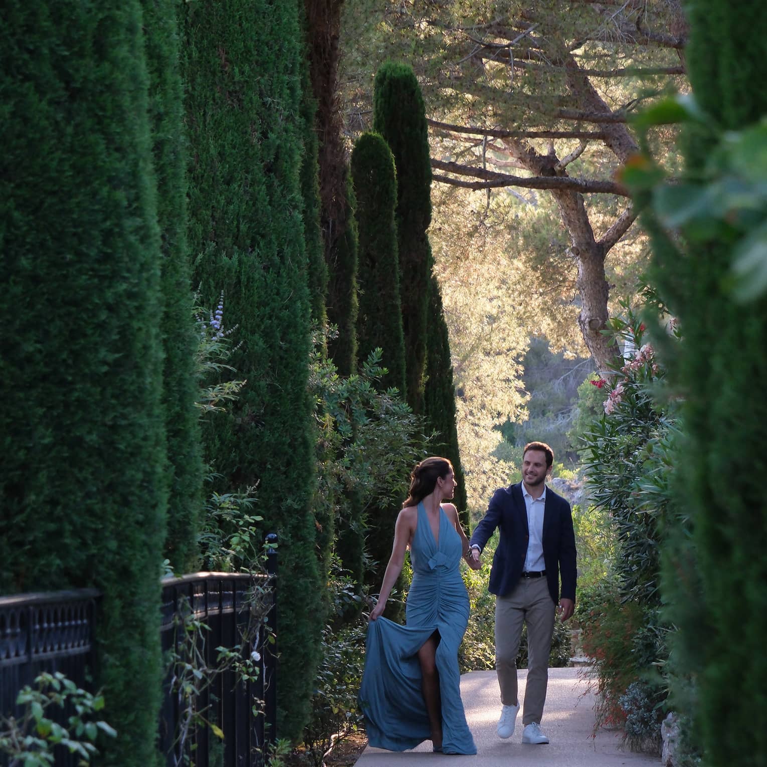A couple dressed in formal attire walks along a shaded pathway surrounded by tall, lush trees