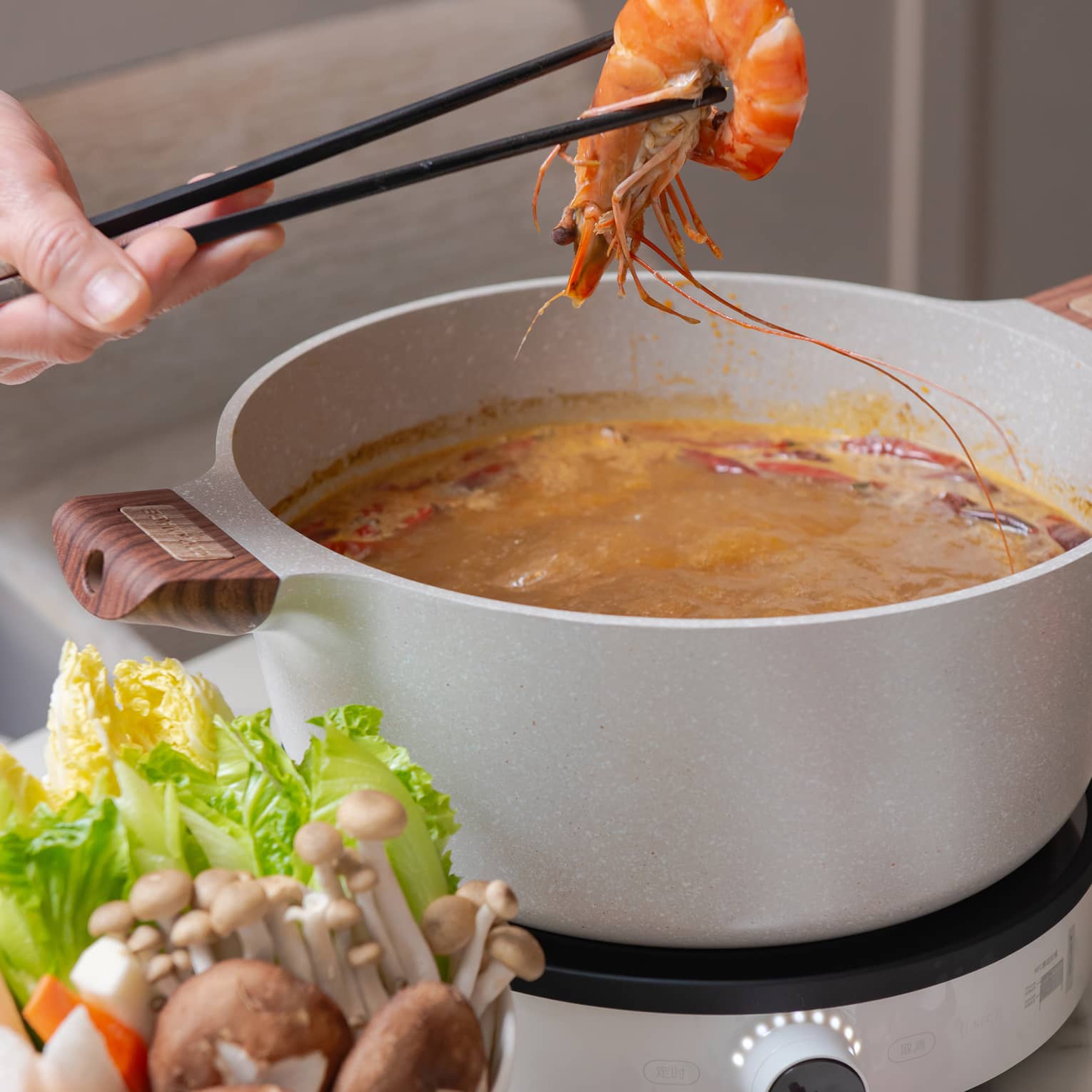 Chopsticks lift a piece of shrimp from a simmering pot on a hotplate, next to a small bowl of mushrooms and lettuce.