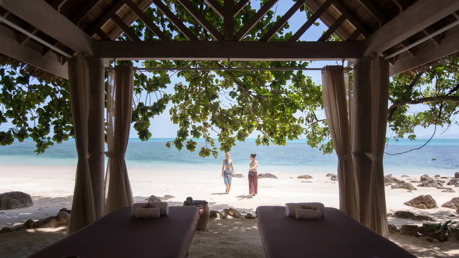 Couples massage beds under cabana in front of two women walking on white sand beach