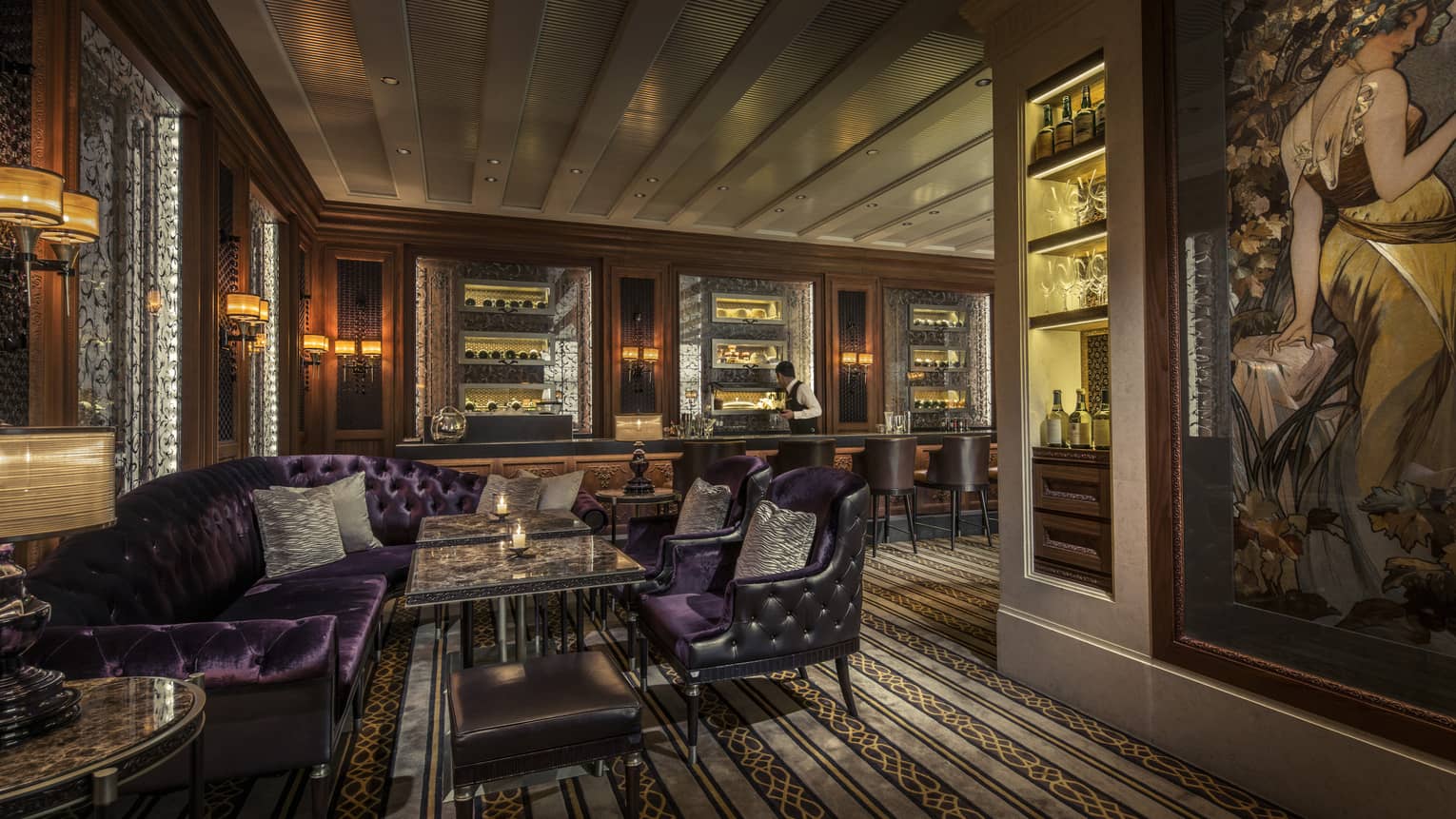 Luxurious lounge with purple velvet seating, marble tables and a bartender preparing drinks behind a backlit bar.