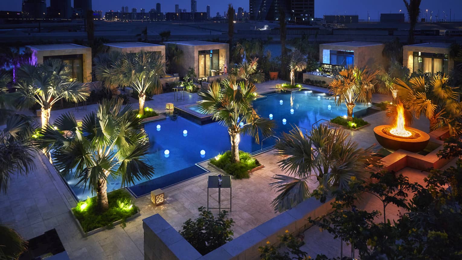 Aerial view of swimming pool, palm trees at night with lights, patio with large outdoor fireplace