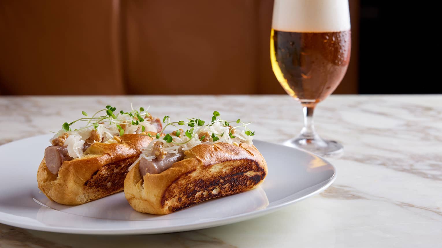 Two Hot Dogs Topped with Sauerkraut and Microgreens on white plate beside dark beer in glass