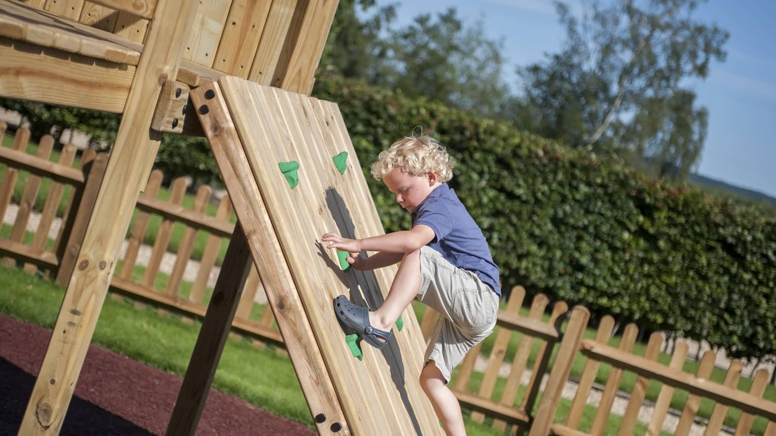 Boy with curly blonde hair scales a wooden climbing wall on outdoor activity set