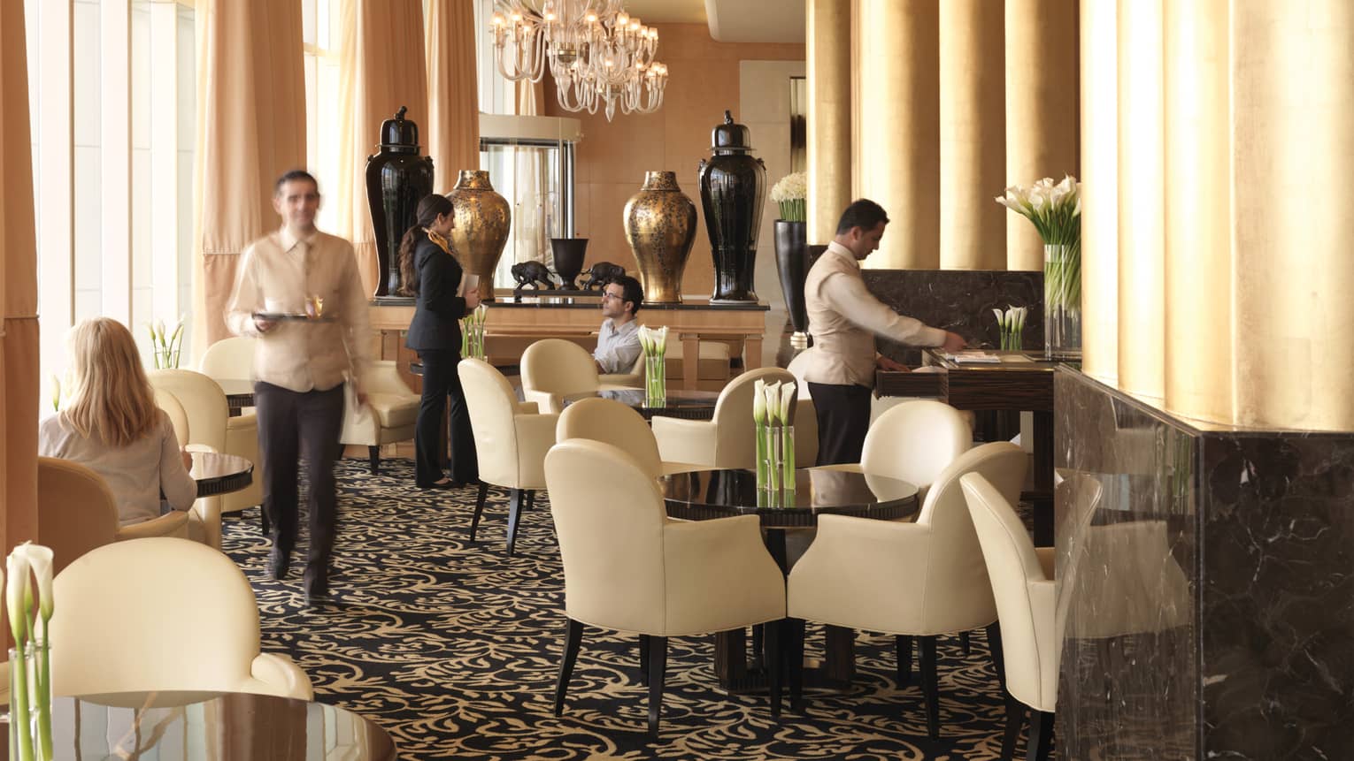 Sunny lobby lounge, hotel staff talk to guests at tables with white leather dining chairs, big gold and black vases behind