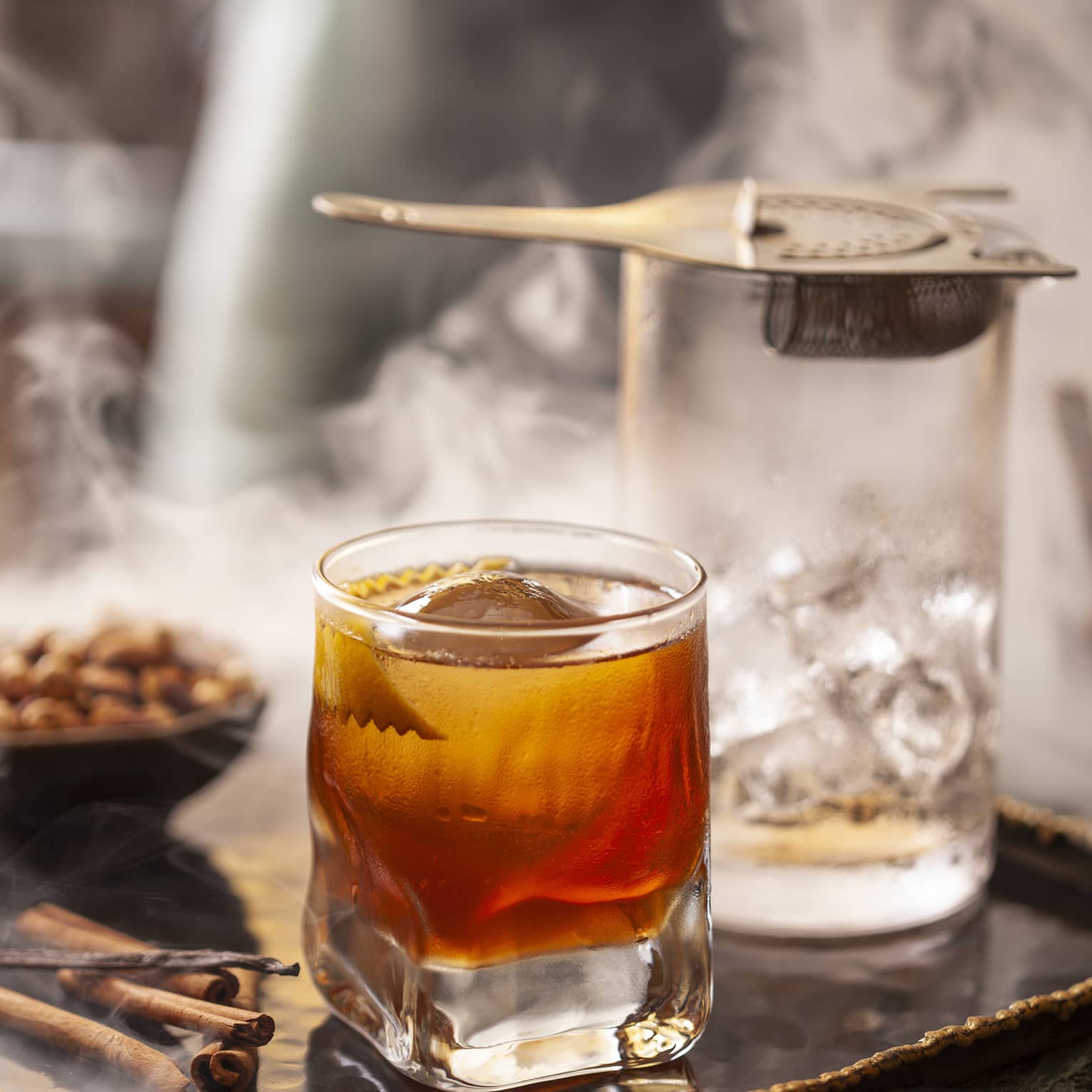 Smoke swirls around an elegant tray holding a glass of bourbon, a glass of ice with a strainer and some cinnamon sticks.