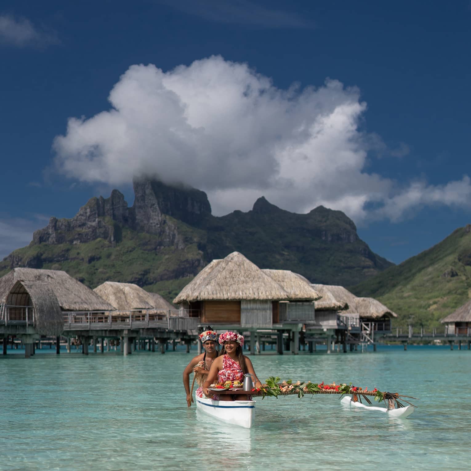 Two vibrantly dressed people in a canoe proffer a tray with colourful food attached to a pole draped with leaves and flowers.