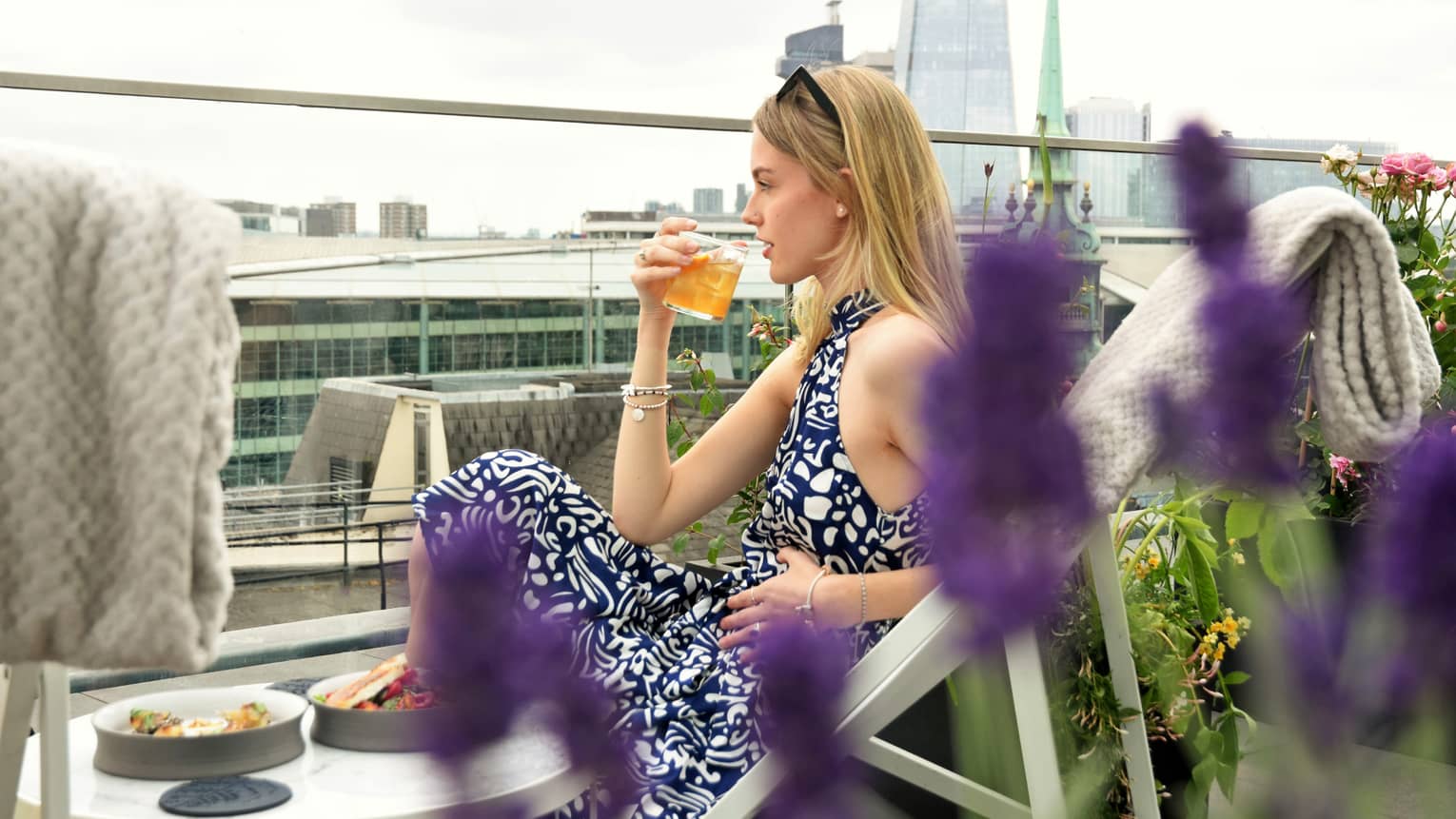 Blonde woman in lounge chair sipping a cocktail on terrace overlooking London