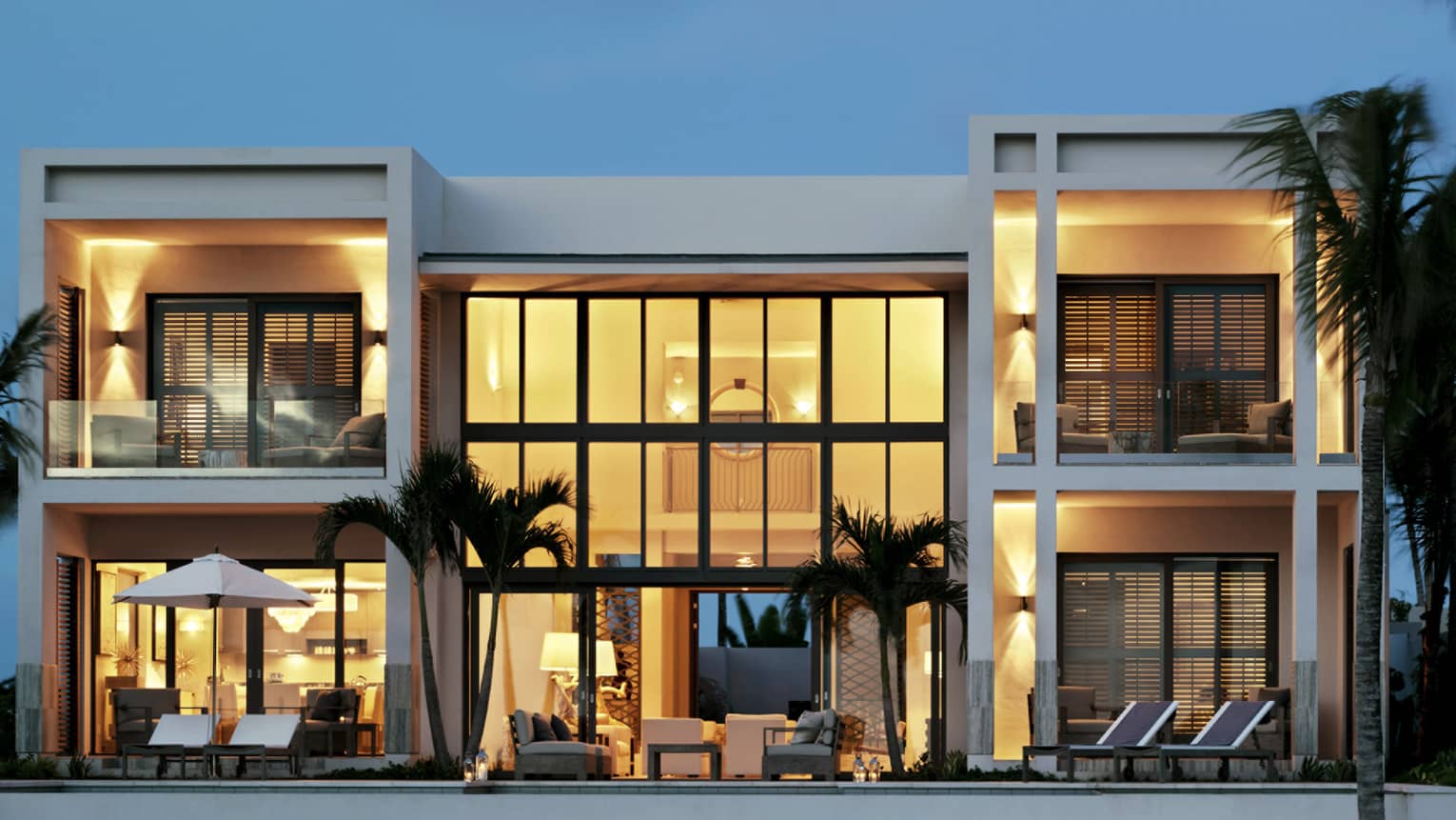 Exterior view of five-bedroom, white beachfront villa at dusk, two storeys with floor-to-ceiling illuminated windows