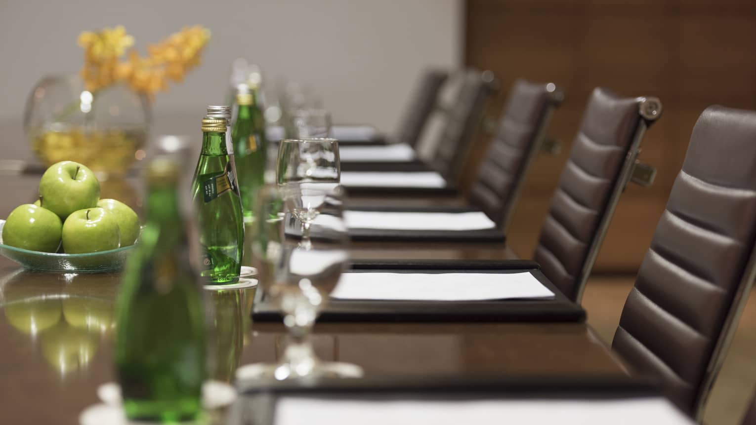 Close-up of business meeting table row of chairs, paper agendas, green glass bottles, apples