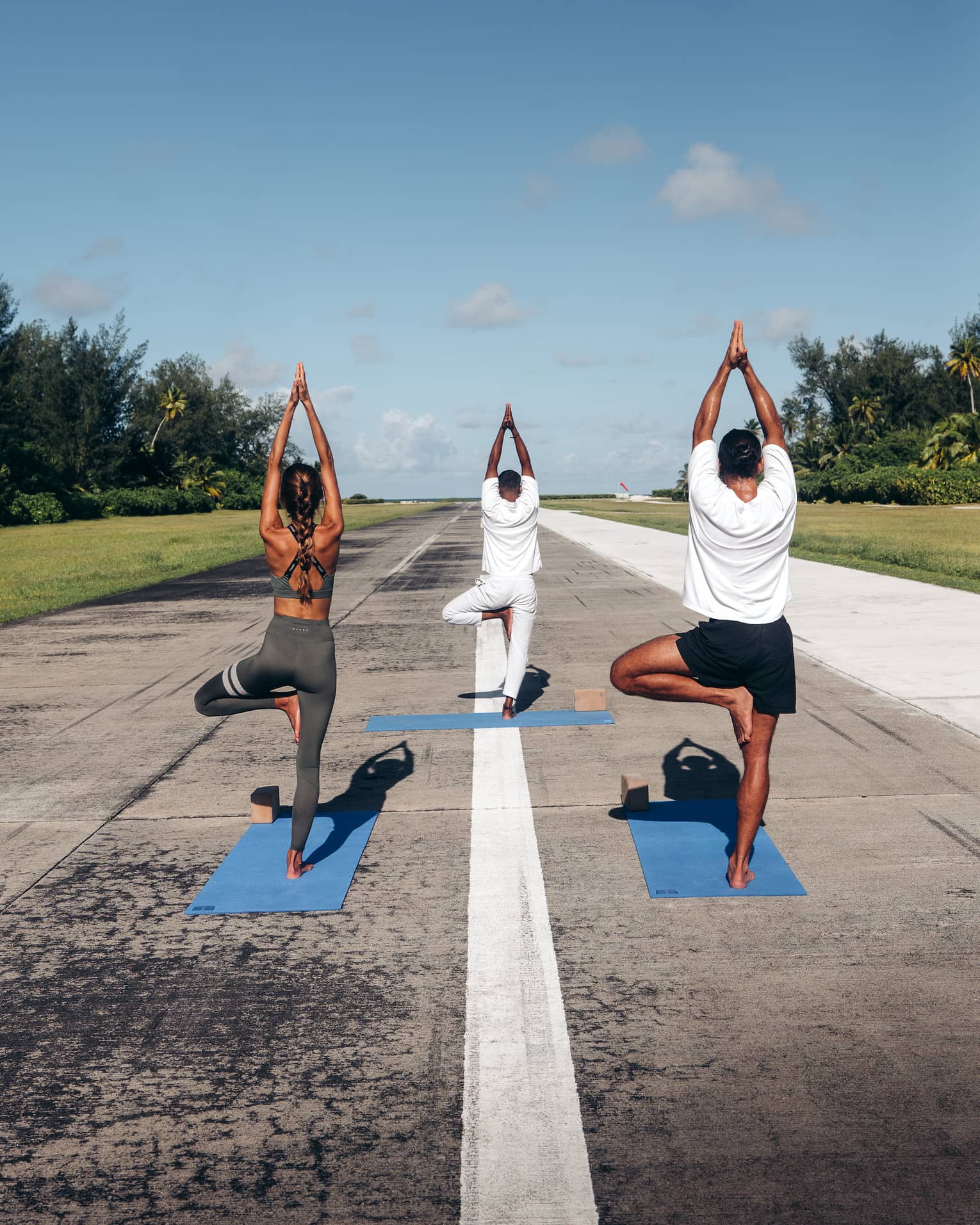 Rear view of three people practicing yoga in tree pose on an airport runway framed by lush greenery, under a clear blue sky.
