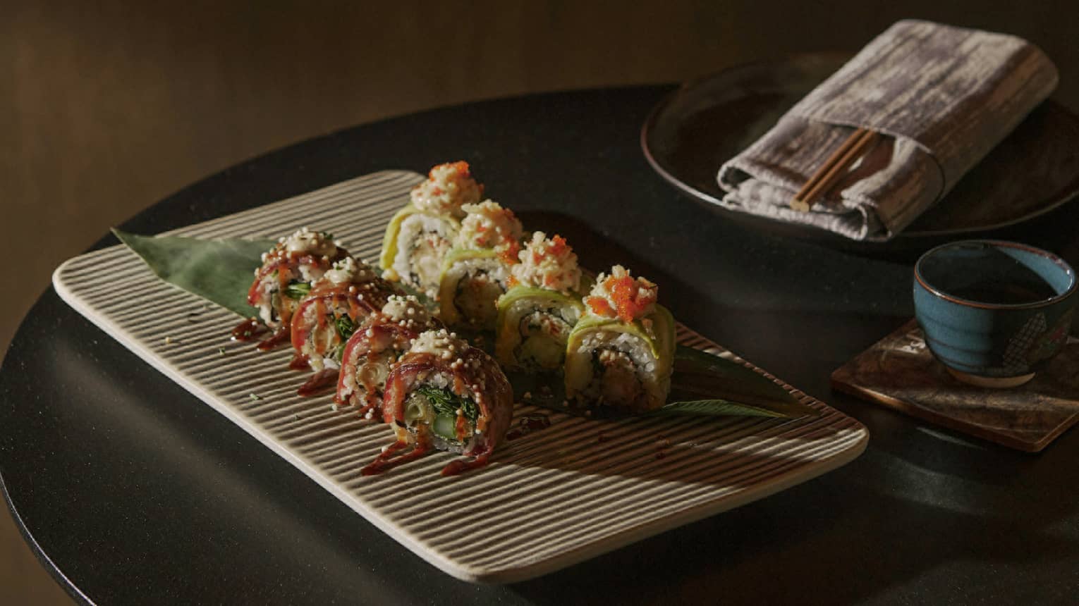 Two rows of four colourful Sushi pieces atop ecru rectangular striped dish beside chopsticks in napkin