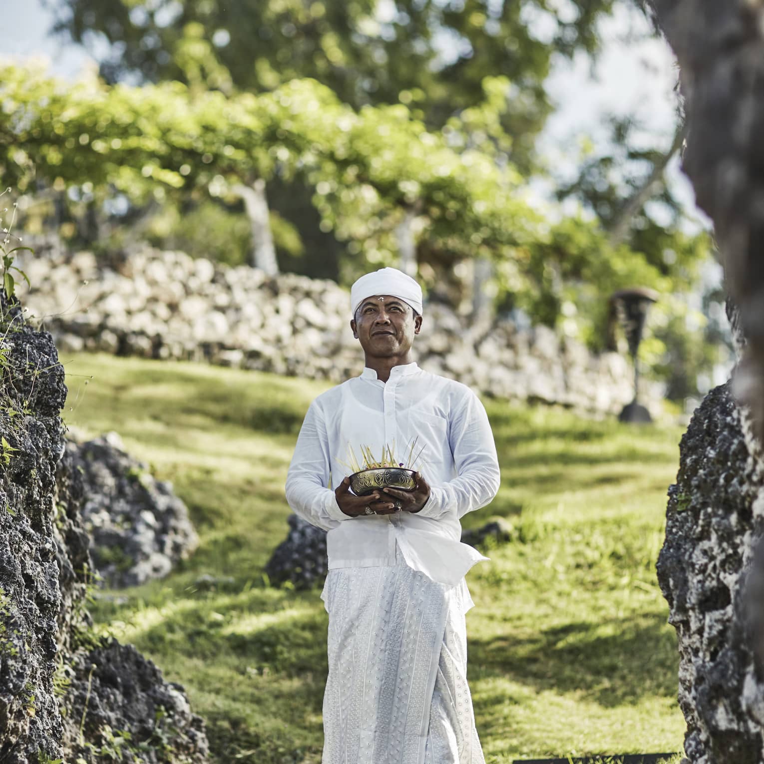 A priest in white attire stands on a stepping stone on a grassy hill, eyes gazing ahead, and holds a bowl of incense sticks.