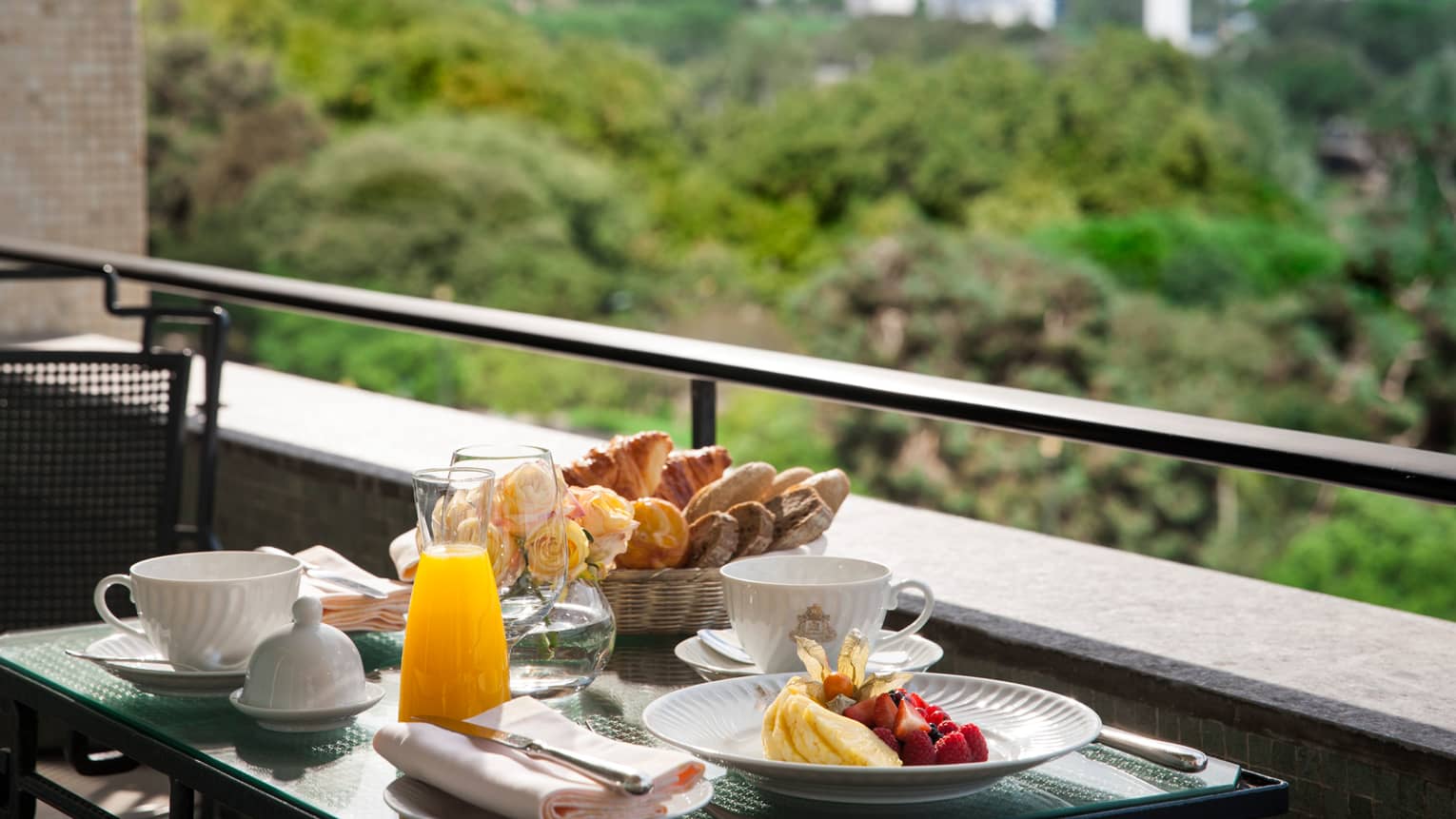 Patio dining table with plate of tropical fruit, glass jug with orange juice, coffee mugs, fresh roses and bread basket