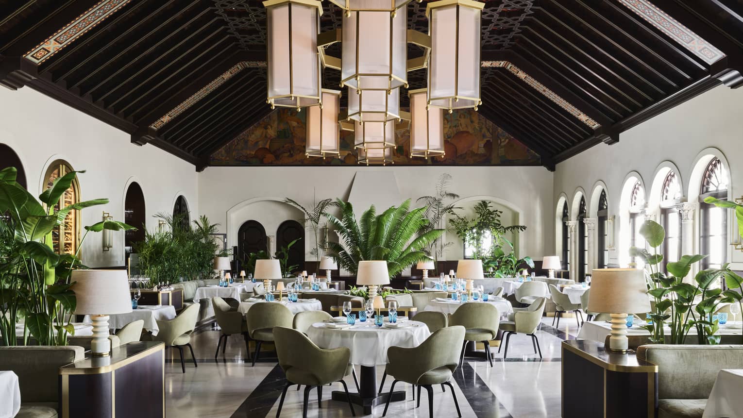A sophisticated dining room with high, dark wood-beamed ceilings, large hanging lantern chandeliers, green upholstered chairs, white tablecloths and indoor plants.