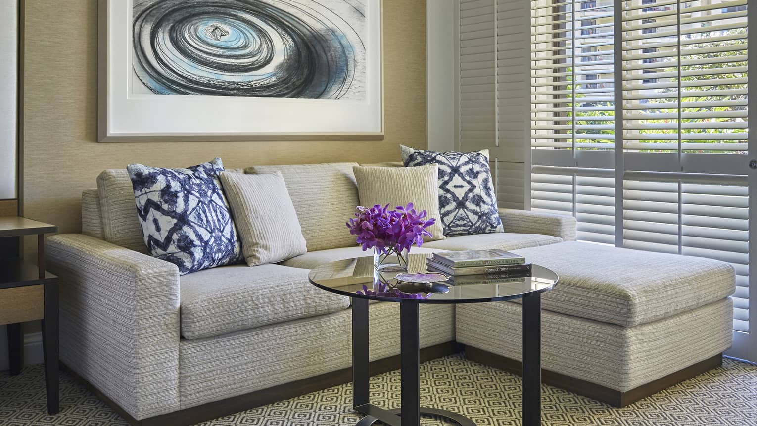 Cozy sitting area with light sectional sofa, patterned pillows, glass coffee table and abstract wall art near window blinds