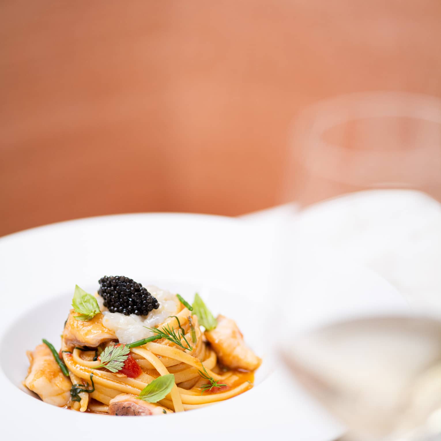 A swirl of seafood linguine topped with black caviar and fresh herbs in a wide pasta bowl, served with a glass of white wine.