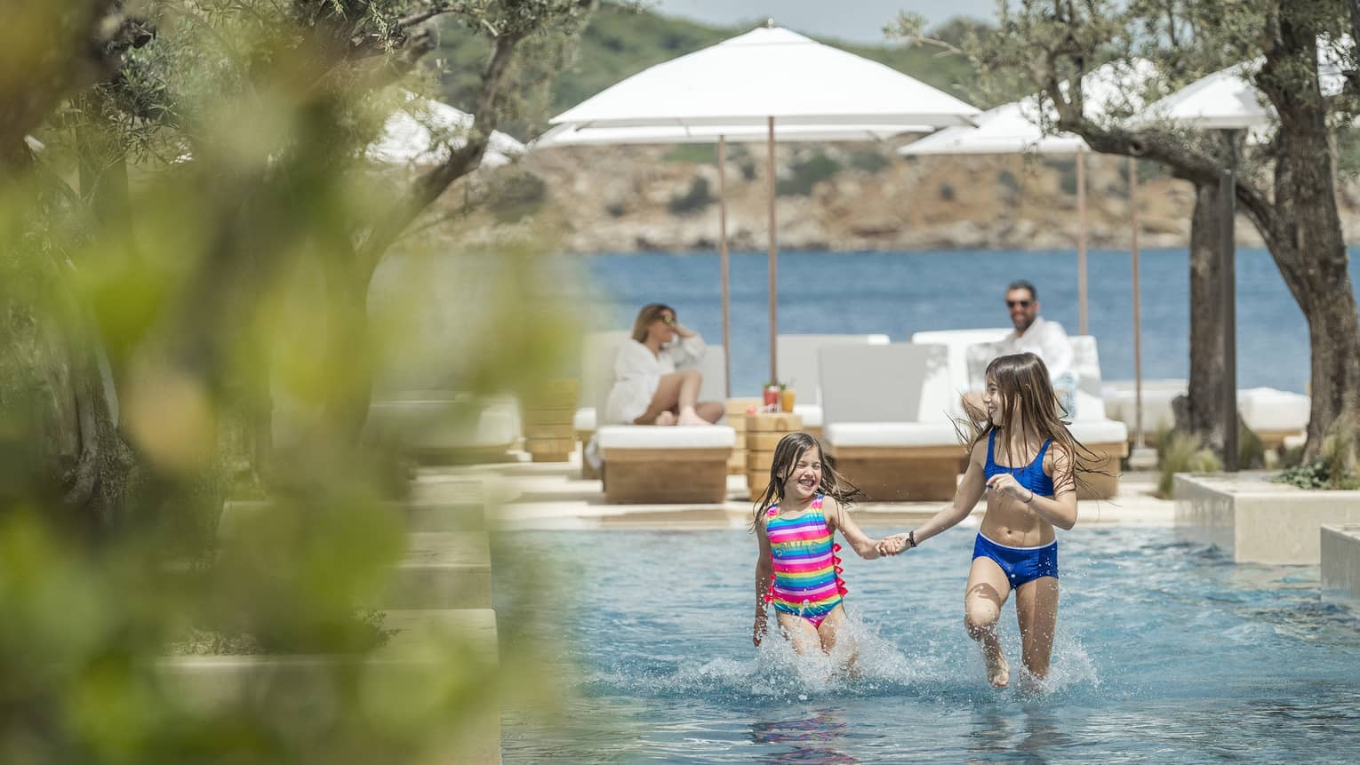 Two smiling children frolick in a shallow outdoor pool while their parents watch from sun loungers. The ocean is behind them.