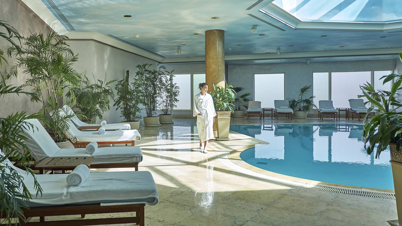 Woman in white bathroom walks the length of the indoor pool with skylight; potted plants and pool longues surrounding