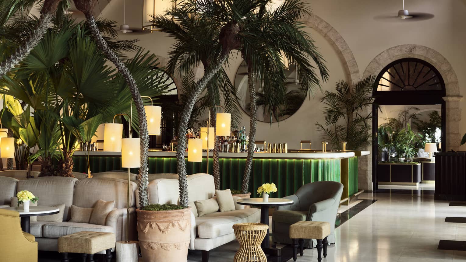 Indoor lounge with palm trees, white upholstered arm chairs, green bar in the background