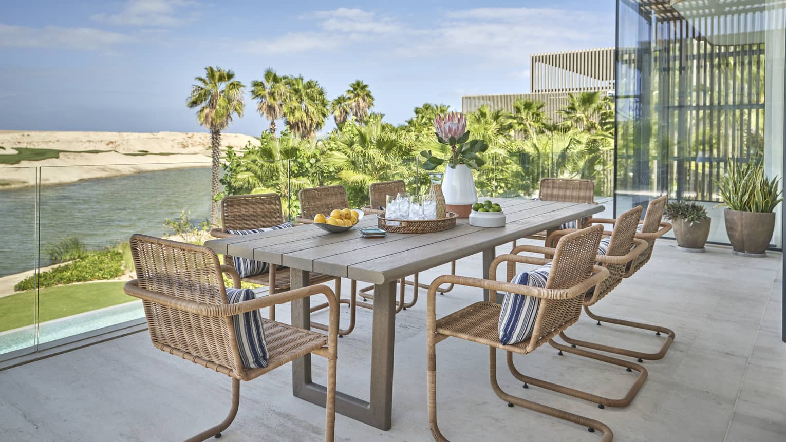 Terrace with rectangular dining table and chairs and glass railing, overlooking water and golf course with palm trees