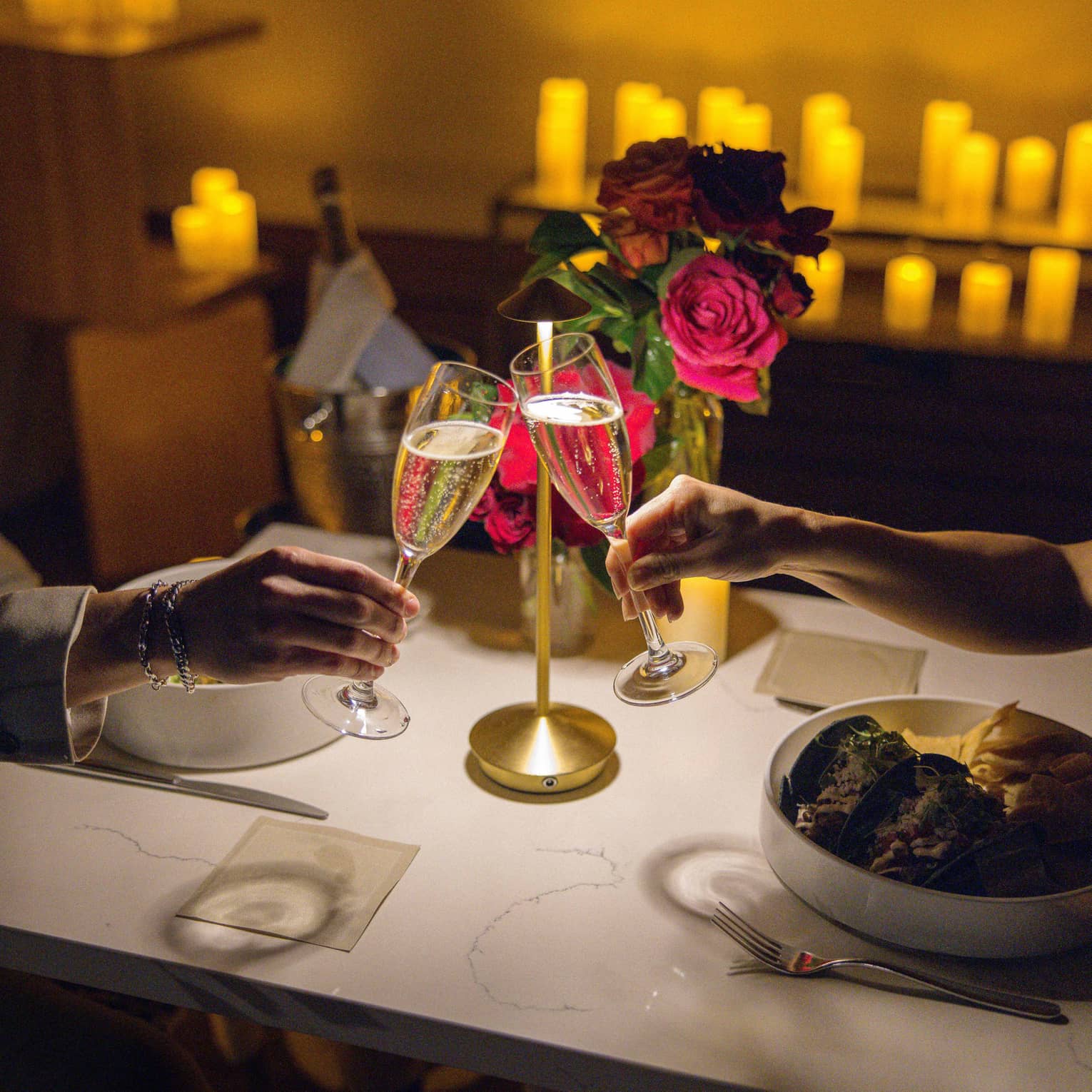Two hands clink champagne glasses over a dining table set with flowers, candles in the background