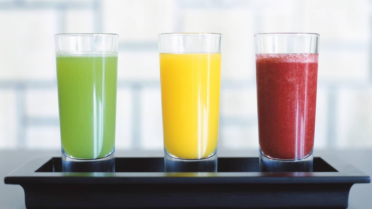 Three tall juice glasses with colourful green, orange and red smoothie drinks on tray